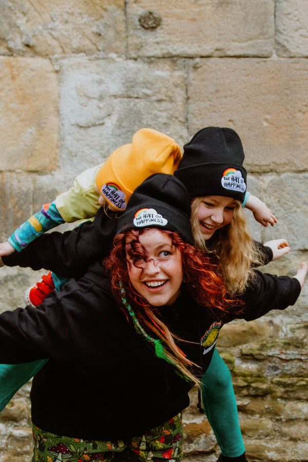 The Hat of Happiness beanie in yellow and black with embroidered lettering on a cloud and rainbow. 100% soft-touch acrylic in a double layer knit. Designed by Katie Abey in the UK