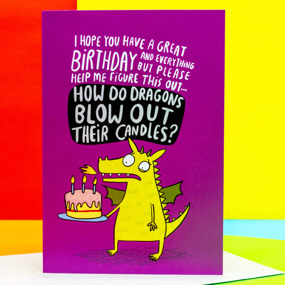 A6 greeting birthday card. Illustrated green dragon holding birthday cake, text reads 'I hope you have a great birthday and everything but please figure this out... how do dragons blow out their candles?' On a pink background. Designed by Katie Abey in the UK