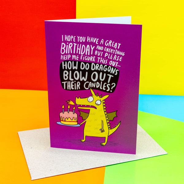 A6 greeting birthday card. Illustrated green dragon holding birthday cake, text reads 'I hope you have a great birthday and everything but please figure this out... how do dragons blow out their candles?' On a pink background. Designed by Katie Abey in the UK
