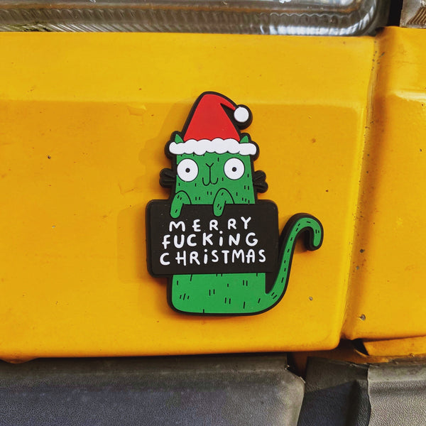 Merry Swear Word green cat fridge magnet holding funny sign and wearing red Santa hat. Designed in the UK by Katie Abey