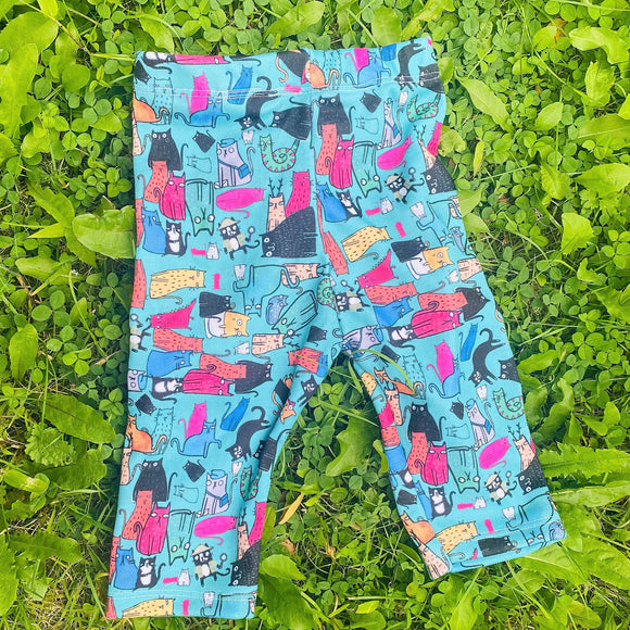All of the cats baby leggings 0 - 3 months old size. The leggings are laying flat on a grassy area. The legging print are a blue base with various repeating cats in different colours, patterns and sizes. Designed by Katie Abey in the UK.