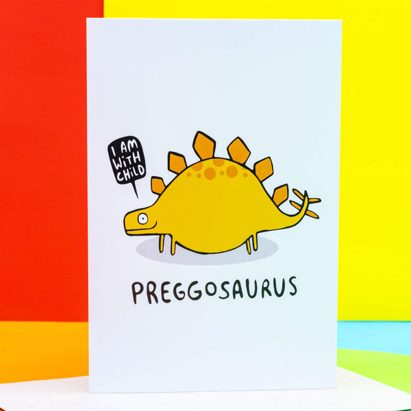 The Preggosaurus Pregnancy a6 Greeting Card designed and printed in the UK by Katie Abey. The card is stood upright on the brown envelope it comes with and sweets surrounding it on a white background. The front cover is a white background with a pregnant yellow stegosaurus smiling with a speech bubble reading 'I am with child' and text underneath them reading 'Preggosaurus'.