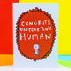 Congrats on your tiny human A6 greeting card designed by Katie Abey and printed in the UK. The card is a white background with a red frilly frame shape with a cartoon white baby and text above that reads 'congrats on your tiny human'. The card is stood up on the brown envelope it comes with on a colourful background.