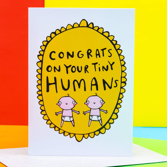 Congrats on your tiny humans A6 greeting card designed by Katie Abey and printed in the UK. The card is a white background with a gold frilly frame shape with two cartoon white babies and text above that reads 'congrats on your tiny humans'. The card is stood up on the brown envelope it comes with on a colourful background.