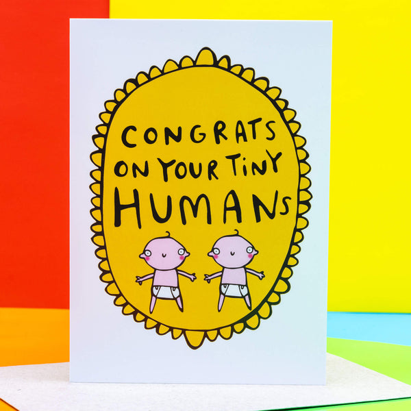 Congrats on your tiny humans A6 greeting card designed by Katie Abey and printed in the UK. The card is a white background with a gold frilly frame shape with two cartoon white babies and text above that reads 'congrats on your tiny humans'. The card is stood up on the brown envelope it comes with on a colourful background.