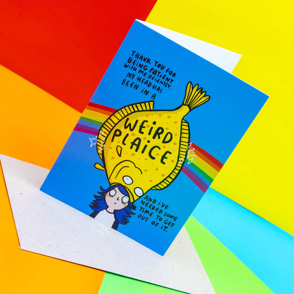 A6 Greetings Postcard reads 'Thank you for being patient with me recently my head has been in a Weird Plaice and I've needed some time to get out of it'. Blue background with rainbow illustration, featuring a yellow plaice fish eating a girls head with blue hair on their head.