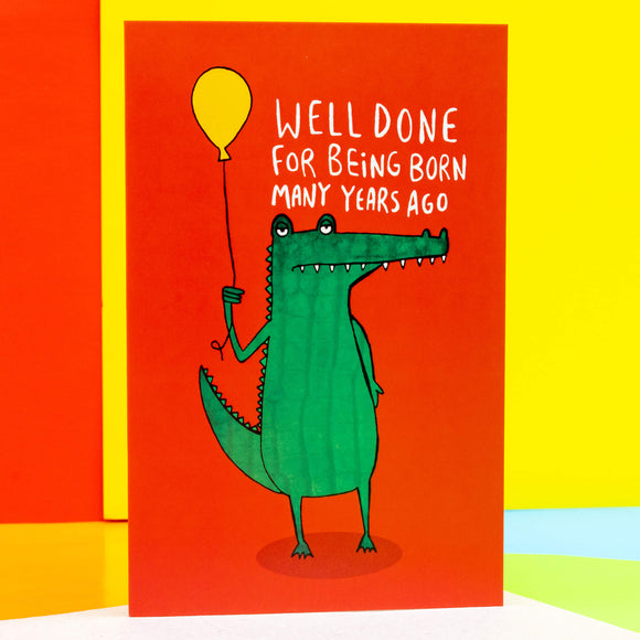 a red A6 greeting card with a illustration of an unimpressed looking green crocodile holding a yellow balloon with white text above it that reads 'well done for being born many years ago'. Designed by Katie Abey in the UK.