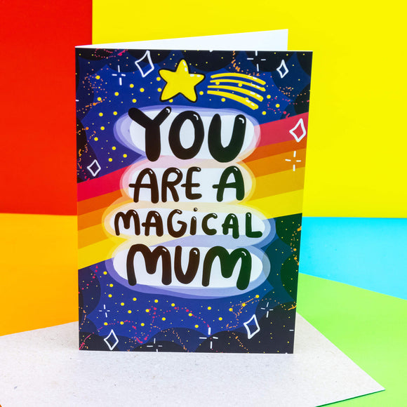 You are a magical mum a6 greeting card stood up on a mossy rock outside. The front cover is a dark blue to black galaxy background with various red, white, yellow stars and sparkles, a red, orange, yellow gradient stripes going across the centre. The centre text reads 'You are a magical mum' with a yellow shooting star above it. Designed by Katie Abey in the UK.