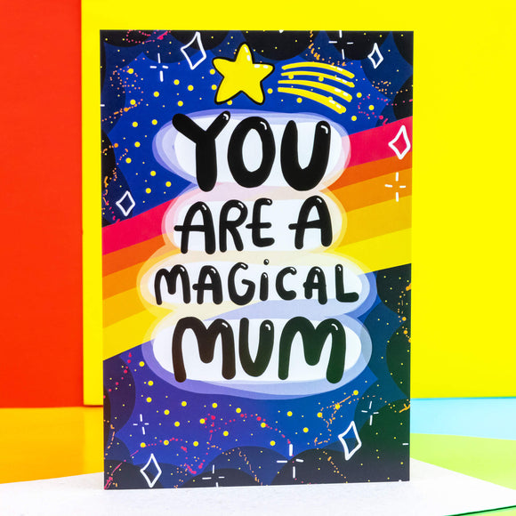 You are a magical mum a6 greeting card stood up on a mossy rock outside. The front cover is a dark blue to black galaxy background with various red, white, yellow stars and sparkles, a red, orange, yellow gradient stripes going across the centre. The centre text reads 'You are a magical mum' with a yellow shooting star above it. Designed by Katie Abey in the UK.